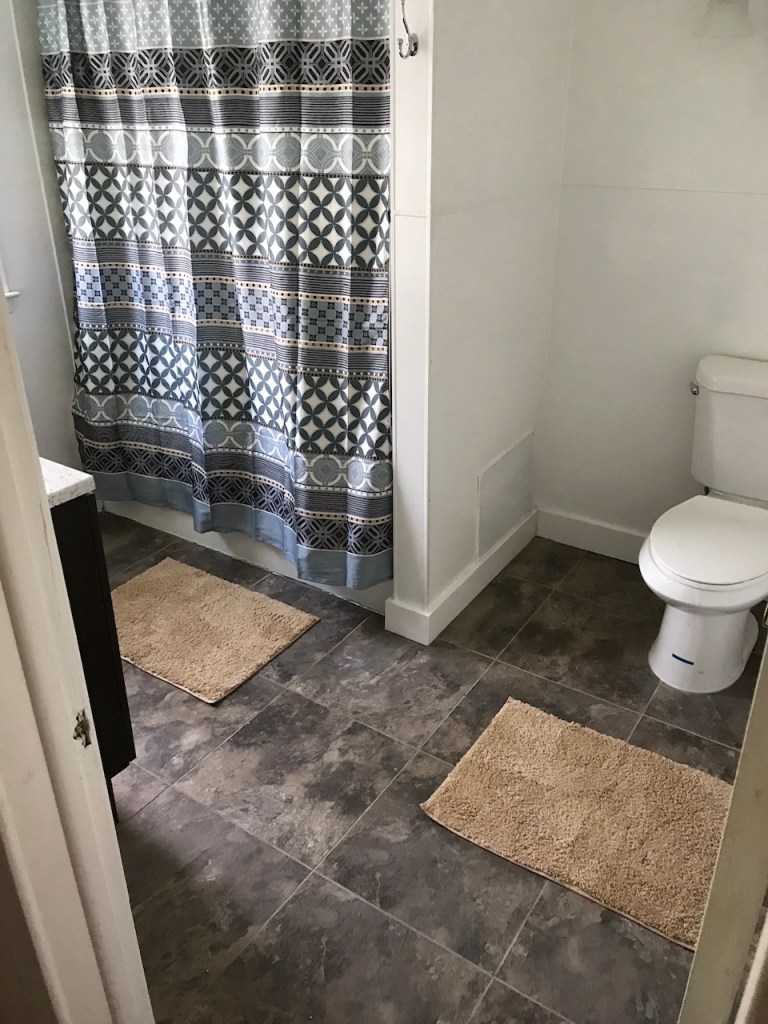 bathroom with toilet. black tile floor with brown mats and blue shower curtain