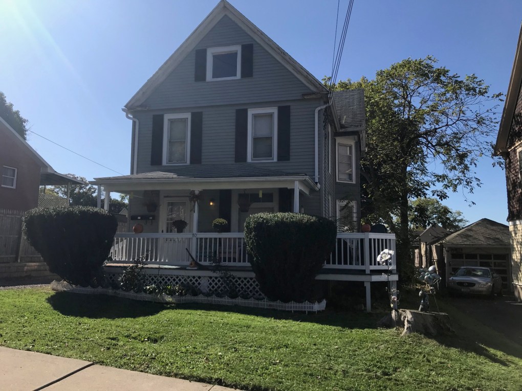 green house with black shutters and white porch with two headges in front of porch