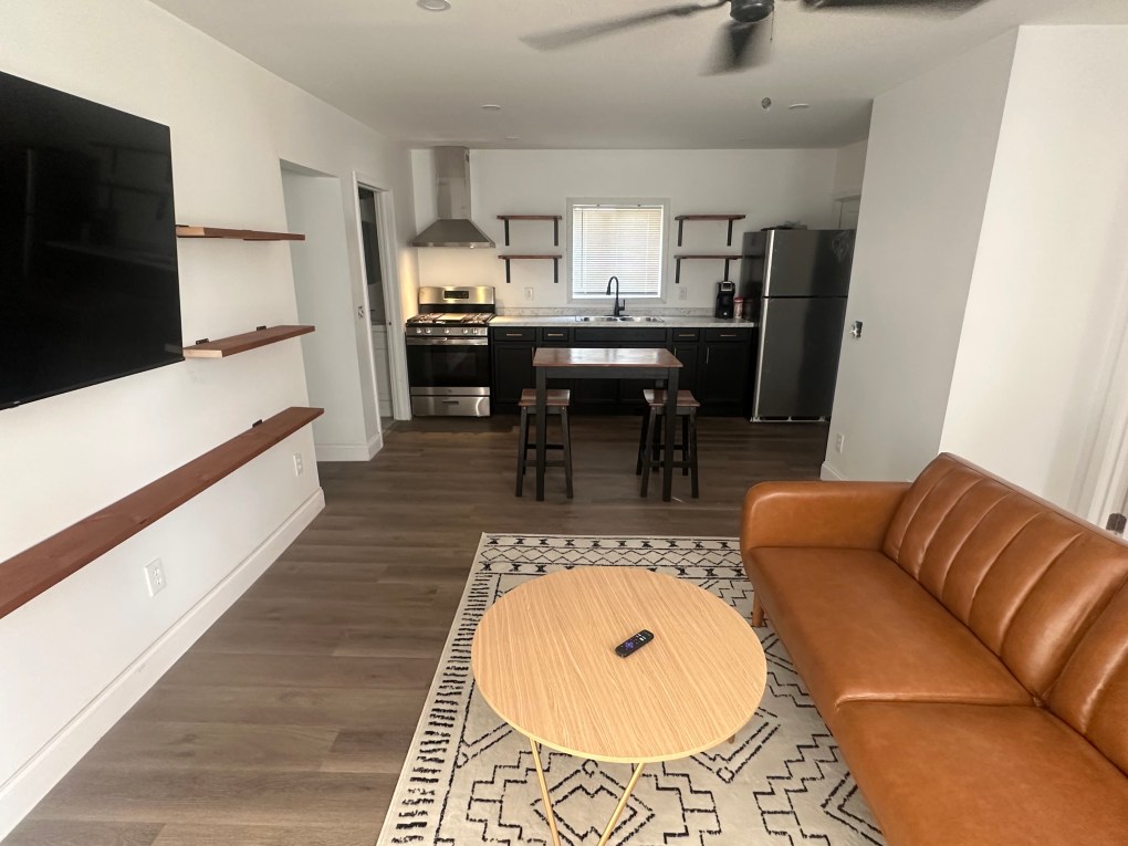 Living room that has brown faux leather couch and light round brown table looking into kitchen that has black cabinets and stainless steel appliances.